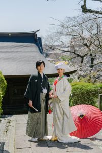 お寺を背景に羽織袴、白無垢で赤い和傘を脇に撮影をした新郎新婦